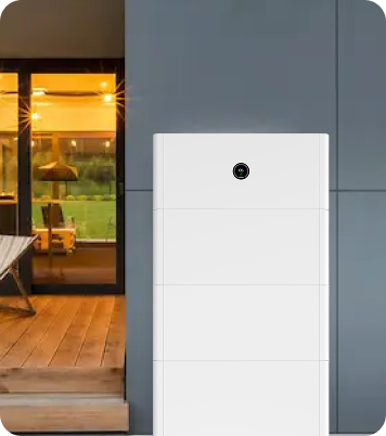 A white home battery storage unit installed on a modern home's exterior wall next to a lit patio