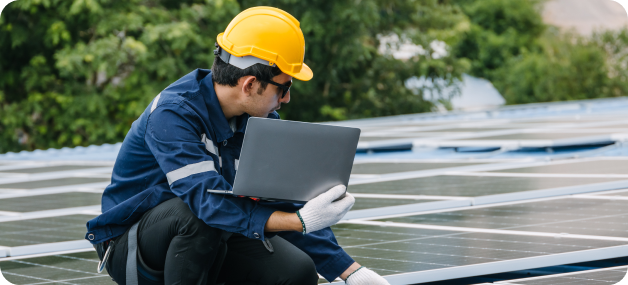 A professional solar technician in a hard hat inspecting solar panels on a roof with a laptop